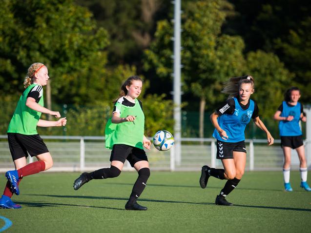 Drei Fußballspielerinnen in Trainingswesten schießen einen Ball auf einem Kunstrasenplatz unter blauem Himmel.