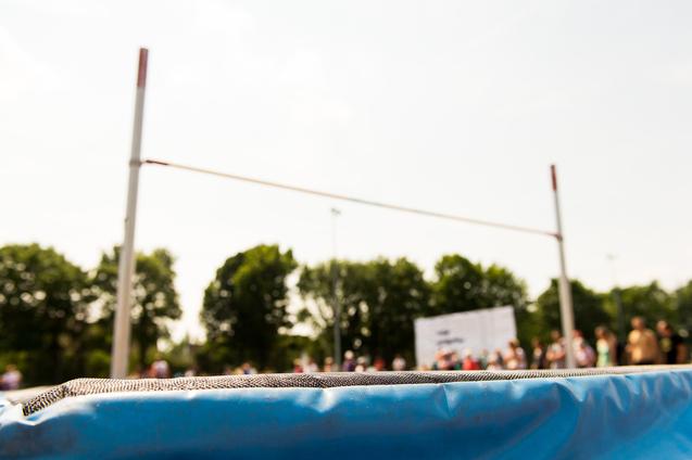 Nahaufnahme einer blauen Sprungmatte mit einer Hochsprunganlage und unscharfem Publikum im Hintergrund.